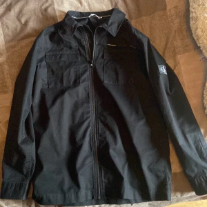 Svart overshirt från Calvin Klein Jeans - Svart overshirt från Calvin Klein Jeans med klassisk krage, två bröstfickor och dragkedja framtill. Snygg CK-logga på ärmen ger en clean och modern vibe. Perfekt för lager-på-lager och enkel att matcha med allt.
