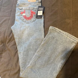 NYA True Religion bootcut jeans ljusblå - Helt NYA blå utsvängda jeans från True Religion med broderad bakficka i rosa och vitt. Klassisk femficksmodell i jeansmaterial och med True Religion-patch bak. Snygg och avslappnad passform. Sitter väldigt snyggt på. Nypris: 1599:- 