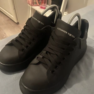 Svarta sneakers Alexander McQueen - Säljer ett par svarta Alexander McQueen sneakers med chunky platt sula och rund tå. Skorna är i skinn med svarta skosnören och diskret logga på plösen och hälen. Klassisk och stilren modell som ger en edgy vibe till outfiten.