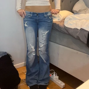 Blå bootcut jeans hål - Säljer ett par ljusblå botcut jeans med låg midja. Baksidan har stora fickor med rosa broderade Juicy-kronor. Jeansen har ljus tvätt, dragkedja och knapp framtill samt coola slitningar på benen. Dessvärre har varan en defekt som ni ser på bild 4 går lätt att sy ihop dock! 