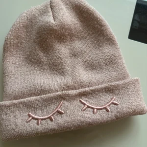 Ljusrosa glittrig mössa H&M barn - Supersöt ljusrosa stickad mössa från H&M med glittriga trådar och broderade ögonfransar framtill. Perfekt accessoar för att hålla värmen och addera lite extra stil till vinterlooken. Passar barn 4-8 år.