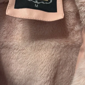 Beige pälsfodrad skinnjacka med bälte - Säljer en snygg beige skinnjacka från Joy i storlek M. Jackan har ett mjukt och fluffigt foder i fuskpäls, stor krage och huva samt bälte i midjan för extra stilpoäng. Perfekt för dig som vill ha en varm och trendig look under kallare dagar.