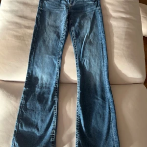 Blå bootcut jeans lågmidjade från Gina Tricot - Snygga blå jeans från Gina Tricot med bootcut passform och klassiska fem fickor. Jeansen har dubbla knappar i midjan, mellanblåtvätt och kontrastsömmar. Perfekta för dig som gillar en avslappnad men trendig look.