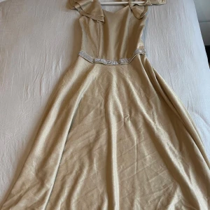 Champagnefärgad långklänning med volang - Säljer en elegant champagnefärgad långklänning med glittrigt tyg och breda volangärmar. Klänningen har smala axelband, v-ringad hals och ett dekorativt band med paljetter i midjan. Perfekt för bal eller fest! Baksidan har smockdetaljer och dragkedja för enkel påklädning.