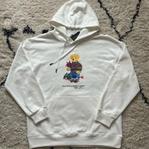 Vit Polo Bear hoodie från Ralph Lauren - Vit hoodie från Ralph Lauren med Polo Bear-tryck på bröstet. Tröjan har huva med snörning, stor magficka och ribbade muddar. Materialet är mjuk bomull och passformen är normal. Perfekt för dig som gillar klassisk streetwear med en twist. Den är för stor, passar inte mig därför säljer jag den.
