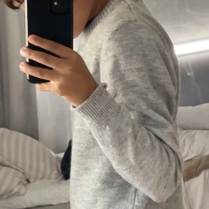 Ljusgrå stickad tröja med rund hals - Säljer en ljusgrå stickad tröja med rund hals och ribbade muddar vid ärmslut och hals. Tröjan har en enkel och clean look som passar perfekt till jeans eller mjukisbyxor. Långärmad och mjuk i materialet för extra komfort.