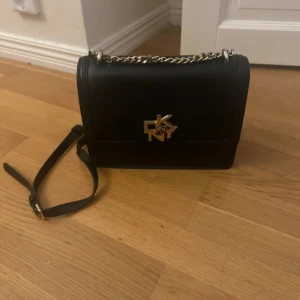 Svart axelväska från DKNY i skinn - Snygg svart axelväska från DKNY med stilren design och guldfärgat DKNY-emblem på framsidan. Väskan har en kedja och justerbar axelrem, samt magnetlås. Perfekt storlek för det viktigaste och tillverkad i slätt skinn.