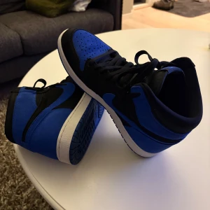 Nike Air Jordan 1 High svart/blå - Säljer ett par Nike Air Jordan 1 High i svart och blått med klassisk siluett. Skorna har svart snörning, blå yttersula och ovandel i mocka och skinn. Perfekta för dig som vill sticka ut med en ikonisk sneaker med snygga kontraster och hög komfort. INGEN SKO LÅDA 