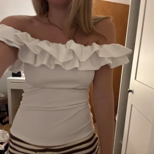 Vit offshoulder volang tubtopp - Supersnygg vit tubtopp med bred volang upptill som går runt hela axelpartiet. Offshoulder och axelbandslös design, sitter tight och har en mjuk, stretchig känsla. Perfekt för dig som vill ha en trendig och feminin look med extra volym upptill.