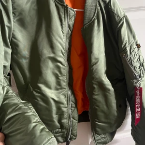 Grön bomberjacka från Alpha Industries - Säljer en klassisk grön bomberjacka från Alpha Industries med orange foder och ikonisk 'Remove Before Flight'-tag på ärmen. Jackan har dragkedja, ribbade muddar och flera fickor, perfekt för en cool streetstil.