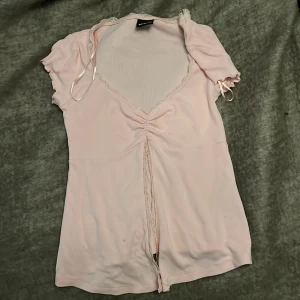 Ljusrosa omlottopp från Gina Tricot - Supersöt ljusrosa omlottopp från Gina Tricot med v-ringning, spetsdetaljer och rynk framtill. Korta ärmar med knyt och mjukt, stretchigt material. Perfekt för en söt och feminin look.