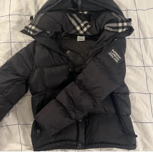 Svart Burberry dunjacka med huva - Säljer en svart dunjacka från Burberry med avtagbara ärmar och huva. Jackan har klassiskt rutigt Burberry-foder i huvan, dragkedja och knappar framtill samt två sidofickor med dragkedja. Perfekt för kalla dagar och riktigt snygg streetstil.