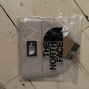 Beige/grå  mössa från The North Face - Snygg beige/gråmössa från The North Face med ribbad struktur och klassisk logga framtill. Perfekt för kalla dagar och enkel att matcha med din stil. Kommer i originalförpackning.