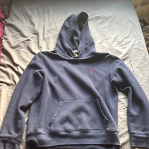 Mörkblå hoodie från Polo Ralph Lauren - Snygg mörkblå hoodie från Polo Ralph Lauren med klassisk röd broderad logga på bröstet. Tröjan har huva, magficka och långa ärmar. Mjuk insida i bomullsmix som gör den extra skön. Perfekt för chill dagar och streetstyle.