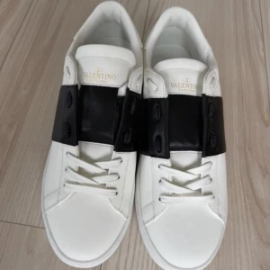 Valentino Garavani Open sneakers - Snygga vita Valentino Garavani Open sneakers med bred svart läderdetalj över snörningen och coola nitar på hälen. Klassisk rund tå och platt sula, tillverkade i skinn med vita skosnören. Perfekt för dig som gillar exklusiv och stilren design.