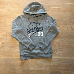 Grå hoodie från Jack & Jones - Snygg grå hoodie från Jack & Jones med tryck på bröstet och klassisk snörning i huvan. Tröjan har långärmade muddar och en mjuk bomullskänsla, perfekt för chill dagar. Stort tryck med 'Authentic' och 'Denim Wear 90' passar runt 170-175
