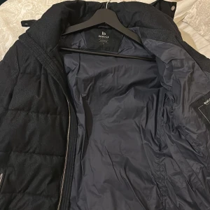 Svart pufferjacka från Boggi Milano - Svart pufferjacka från Boggi Milano med tekniskt tyg, vattenavvisande yta och värmereglering. Jackan har quiltad design, två fickor med dragkedja och hög krage med tryckknappar. Perfekt för kalla dagar och snygg till streetwear.