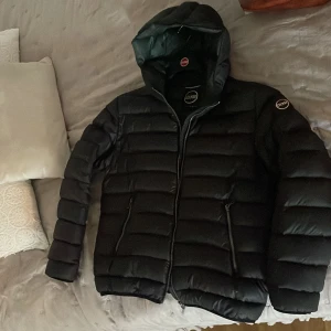 Svart dunjacka från Colmar - Säljer en svart dunjacka från Colmar med huva och dragkedja framtill. Jackan har quiltad design, två fickor med dragkedja och en logga på ärmen. Perfekt för kyliga dagar och riktigt snygg passform. Billigare vid snabb och smidig affär 