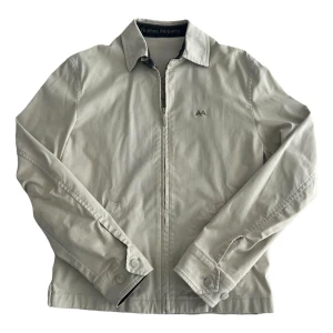 Beige jacka från Thomas Burberry - Stilren beige jacka från Thomas Burberry med klassisk krage, dragkedja framtill och diskret broderad logga på bröstet. Jackan har raka ärmar med knappdetaljer vid mudden och två snedställda fickor. Perfekt för dig som gillar en clean och tidlös look.