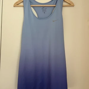Nike Dri-Fit träningslinne - Nike Dri-Fit linne i snygg blå/lila ombre med racerback och diskret logga framtill. Ficka med dragkedja. Storlek M. Använd fåtal gånger, mycket fint skick. Tillverkad i ett lätt, syntetiskt material som andas. Perfekt för träning och sportiga aktiviteter. 
