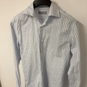 Randig skjorta från Ermenegildo Zegna - Snygg skjorta från Ermenegildo Zegna i storlek S. Den har ett klassiskt randigt mönster i ljusblått och vitt, med krage och knappar framtill. Skjortan är långärmad och tillverkad i mjuk bomull som känns skön mot huden. Perfekt för dig som gillar stilrena och fräscha plagg.