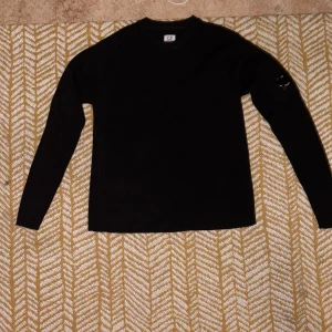 Svart stickad tröja från C.P. Company - Svart stickad tröja från C.P. Company med märket på ärmen. Tröjan har en clean och stilren look. Perfekt för dig som gillar streetwear och vill ha något klassiskt men ändå unikt.