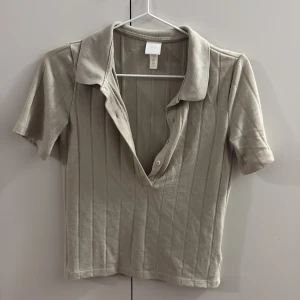 Beige ribbad topp från H&M - Säljer en beige ribbad topp från H&M med korta ärmar och djup v-ringning med knappar. Toppen har en klassisk krage och är tillverkad i mjuk bomullsmix. Perfekt för en stilren och avslappnad look.