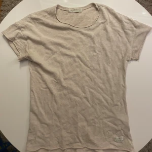 Beige t-shirt från I dig denim - Snygg beige t-shirt från I dig denim med rund hals och korta ärmar. T-shirten är tillverkad i mjuk bomull och har en enkel, clean look med en liten logolapp nedtill. Perfekt för dig som gillar stilrena och bekväma plagg.