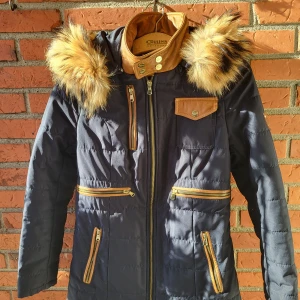 Mörkblå pufferjacka med fuskpäls från Ventiuno - Snygg mörkblå pufferjacka från Ventiuno med bruna detaljer i skinnimitation, guldfärgade knappar och dragkedjor. Jackan har huva med fluffig fuskpäls, flera fickor med dragkedja och hög krage. Perfekt för kalla dagar och riktigt stilren look.