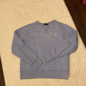 Ljusblå sweatshirt från Polo Ralph Lauren - Snygg ljusblå sweatshirt från Polo Ralph Lauren med klassisk rund hals och broderad vit logga på bröstet. Tröjan har ribbade muddar vid ärmslut och nederkant samt långärmad passform. Perfekt för chill dagar och enkel att matcha med jeans.