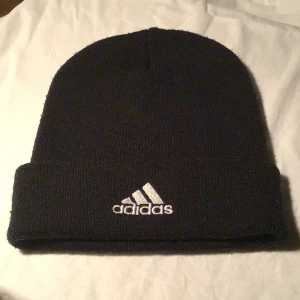 Svart Adidas mössa med broderad logga - Svart mössa från Adidas med klassisk broderad logga i vitt framtill. Mössan är stickad och har en bred uppvikt kant för extra stilpoäng. Perfekt för kalla dagar och enkel att matcha med din streetwear eller sportiga look.