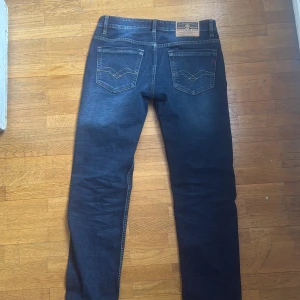 Replay Blue Jeans mörkblå jeans 30/32 - Snygga mörkblå Replay Blue Jeans med klassisk femficksdesign och kontrastsömmar. Märket syns med röd broderad Replay-logga vid fickan. Jeansen har rak passform och är tillverkade i slitstarkt denim-material. Perfekt för dig som gillar stilrena och tidlösa jeans.