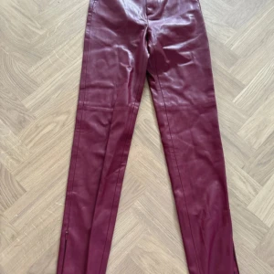 Vinröda skinnbyxor från Zara XS - Snygga vinröda skinnbyxor från Zara i storlek XS. Byxorna har raka ben, hög midja och dragkedjor vid bensluten för extra edge. Materialet är mjukt syntetskinn med en matt finish och coola sömdetaljer längs benen.