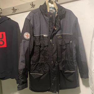 Svart parkajacka från Mountain Horse - Parkajacka från Mountain Horse i svart med mörkgrå detaljer och reflexband. Jackan har flera fickor med lock, dragkedja och knappar framtill samt broderad patch på ärmen. Fodret är ljusgrått med logomönster. Perfekt för dig som gillar outdoor och ridning.