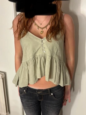 Ljusgrön blus med volang och knappar - Säljer en ljusgrön blus med broderade blommor, volang nedtill och v-ringad hals. Blusen har knappar framtill och är ärmlös, perfekt för varmare dagar. Croppad passform som ger en avslappnad och trendig look.