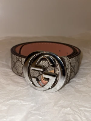 Bälte med silverdetalj - Säljer ett bälte med stor silverfärgad logga som spänne. Bältet har klassiskt beige och brunt mönster med signaturtryck och är tillverkat i skinn med snygga sömmar. Perfekt accessoar för att lyfta din outfit.