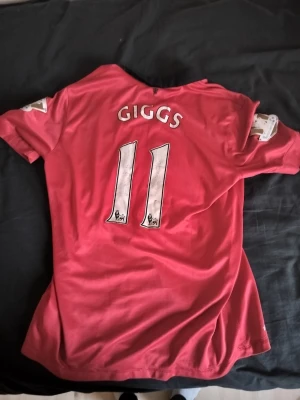 Manchester United tröja Giggs #11 Nike - Säljer en röd Manchester United matchtröja med svart krage, officiellt klubbmärke och AON-tryck på bröstet. På ryggen står det 'Giggs' och nummer 11. Tröjan har knappar vid halsen och är tillverkad i Dri-FIT material.