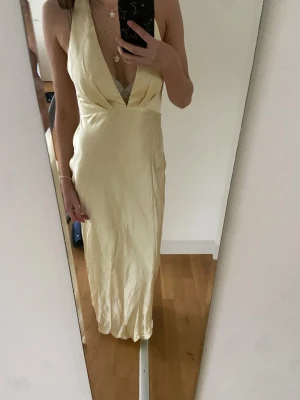 Champagnefärgad långklänning i satin - Säljer en elegant champagnefärgad långklänning med djup v-ringning och halterneck. Klänningen har en slimmad passform och är i satinmaterial med öppen rygg 