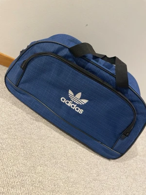 Träningsväska Adidas - Säljer en blå träningsväska från Adidas med vitt logotryck på framsidan. Väskan har ett diskret rutigt mönster, svarta handtag och dragkedjor. Perfekt storlek för gymmet eller sportaktiviteter, med rymligt huvudfack och extra ficka framtill. Perfekt för dig som vill ha plats för sin idrottsutrustning i. Pris går att diskutera 