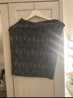 Glittrig svart kortkjol med mönster - Säljer en svart kortkjol med glittrigt geometriskt mönster i silver. Kjolen har en tight passform och är perfekt för dig som gillar att sticka ut. Materialet är mjukt och stretchigt syntet, vilket gör den bekväm att bära. Snygg att matcha med en enkel topp.