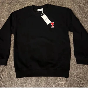 Svart sweatshirt från Ami Paris - Svart sweatshirt från Ami Paris med klassisk rund hals och lång ärm. På bröstet finns ett rött hjärta med bokstaven A. Tröjan är tillverkad i mjuk bomull och har ribbade muddar vid ärmslut och nederkant. Perfekt för en chill och stilren look.