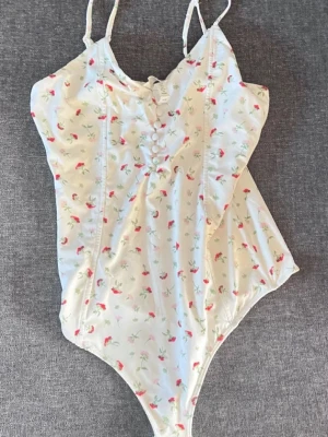 Vit blommig body - Supersöt vit body med små röda och gröna blommor, smala axelband och dekorativa knappar framtill. Perfekt för en somrig look och lätt att matcha med jeans eller kjol. storlek S!!