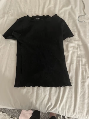 Svart ribbad t-shirt från Monki - Svart t-shirt från Monki med ribbad struktur och vågiga kanter vid halsringning, ärmslut och nederkant. Modellen är kortärmad och har en slim passform som sitter snyggt mot kroppen. Perfekt för dig som gillar minimalistisk stil med en twist.