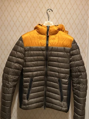 Parajumpers pufferjacka gul/grå - Säljer en Parajumpers pufferjacka i grått och gult med huva och svarta detaljer. Jackan har horisontella quiltade partier, två fickor med dragkedja och en snygg patch på ärmen. Perfekt för dig som vill ha en cool och funktionell jacka.