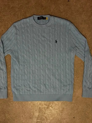 Polo Ralph Lauren Vinter tröja - Kabelstickad ljusblå tröja från Polo Ralph Lauren med klassisk rund halsringning och broderad logga på bröstet. Tröjan har ribbade muddar vid ärmslut och nederkant samt lång ärm. Perfekt för dig som gillar preppy stil.