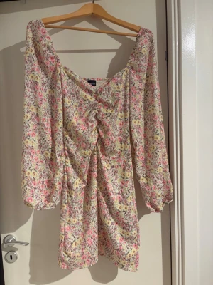 Blommig kortklänning från Gina Tricot - Superfin kortklänning från Gina Tricot med blommigt mönster i ljusa toner av rosa, gul och vit. Klänningen har långärmade puffärmar, v-ringad hals och snygg snörning framtill. Perfekt för sommarens alla tillfällen!