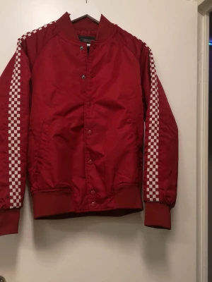 Röd bomberjacka med rutiga detaljer - Röd bomberjacka från 21MEN med vita rutiga ränder längs ärmarna. Jackan har ribbade muddar, tryckknappar framtill och två sidofickor. Materialet är polyester och jackan har en klassisk bomberkrage. Perfekt för dig som vill sticka ut med en sportig vibe.