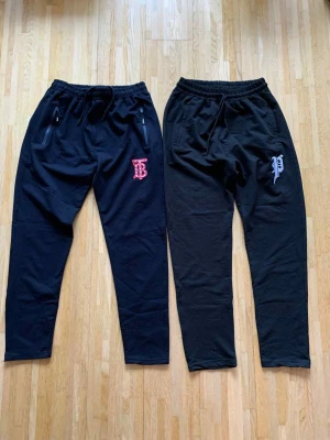 Svarta mjukisbyxor med tryck - Två par svarta mjukisbyxor med resår och snörning i midjan. Ena paret har ett rött tryck med bokstäver på vänster lår och dragkedjor vid fickorna, det andra paret har ett vitt tryck med bokstav och stjärna på vänster lår. Båda har avslappnad passform och är gjorda i mjukt bomullsmaterial.