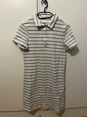 Randig kortklänning från Mango - Säljer en vit kortklänning från Mango med blå horisontella ränder. Klänningen har krage, korta ärmar och knappar framtill. Materialet är mjukt och frottéliknande. Säljer pga. Fel storlek. 