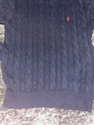 Mörkblå kabelstickad tröja Polo Ralph Lauren - Kabelstickad mörkblå tröja från Polo Ralph Lauren med klassisk röd logga broderad på bröstet. Tröjan har rund halsringning och ribbade muddar. Perfekt för dig som gillar preppy stil och vill ha något både snyggt och bekvämt. HÖR AV ER VID FUNDERINGAR!